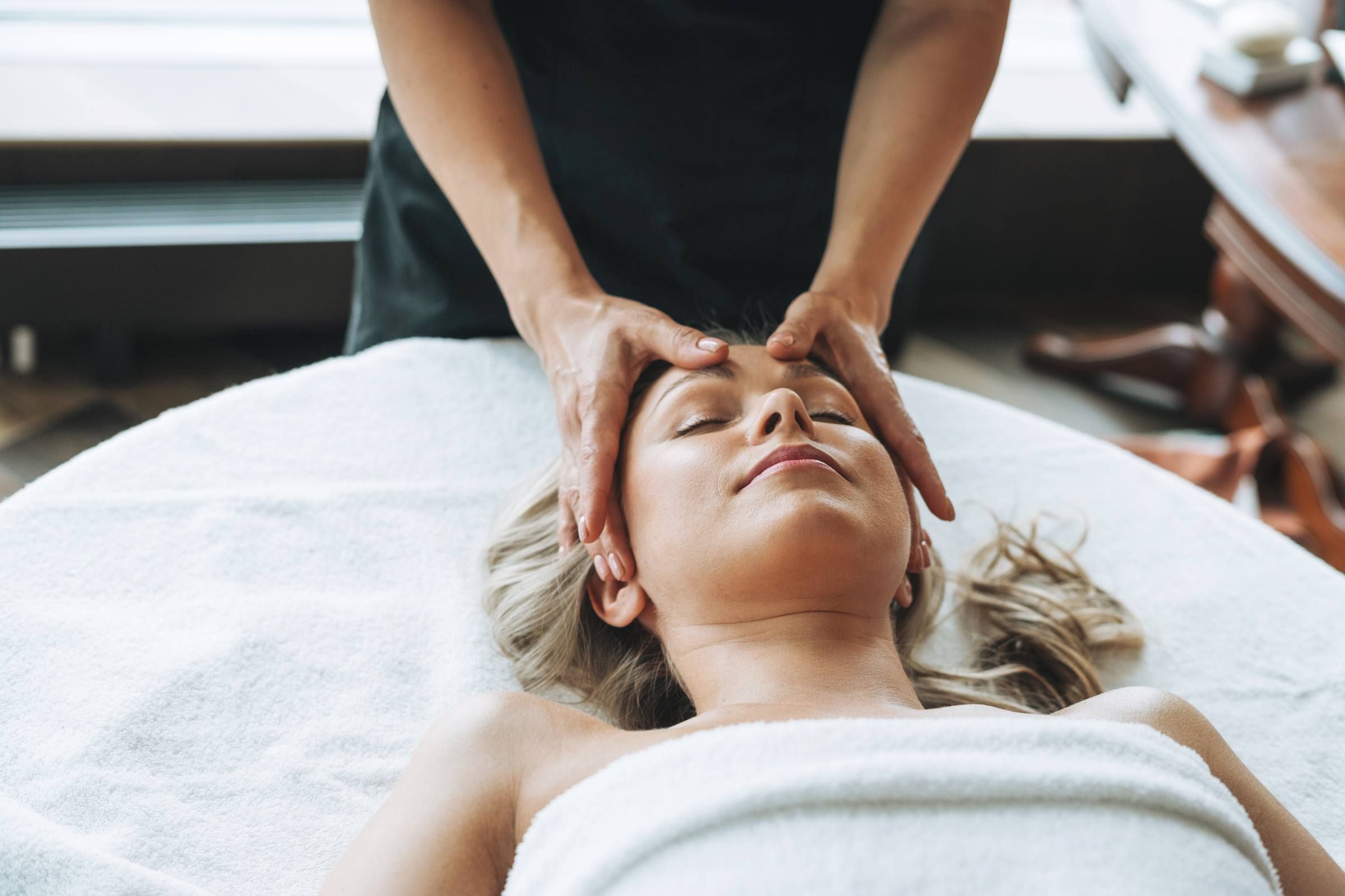 Blonde young woman on couch with white linen enjoys facial massage in spa